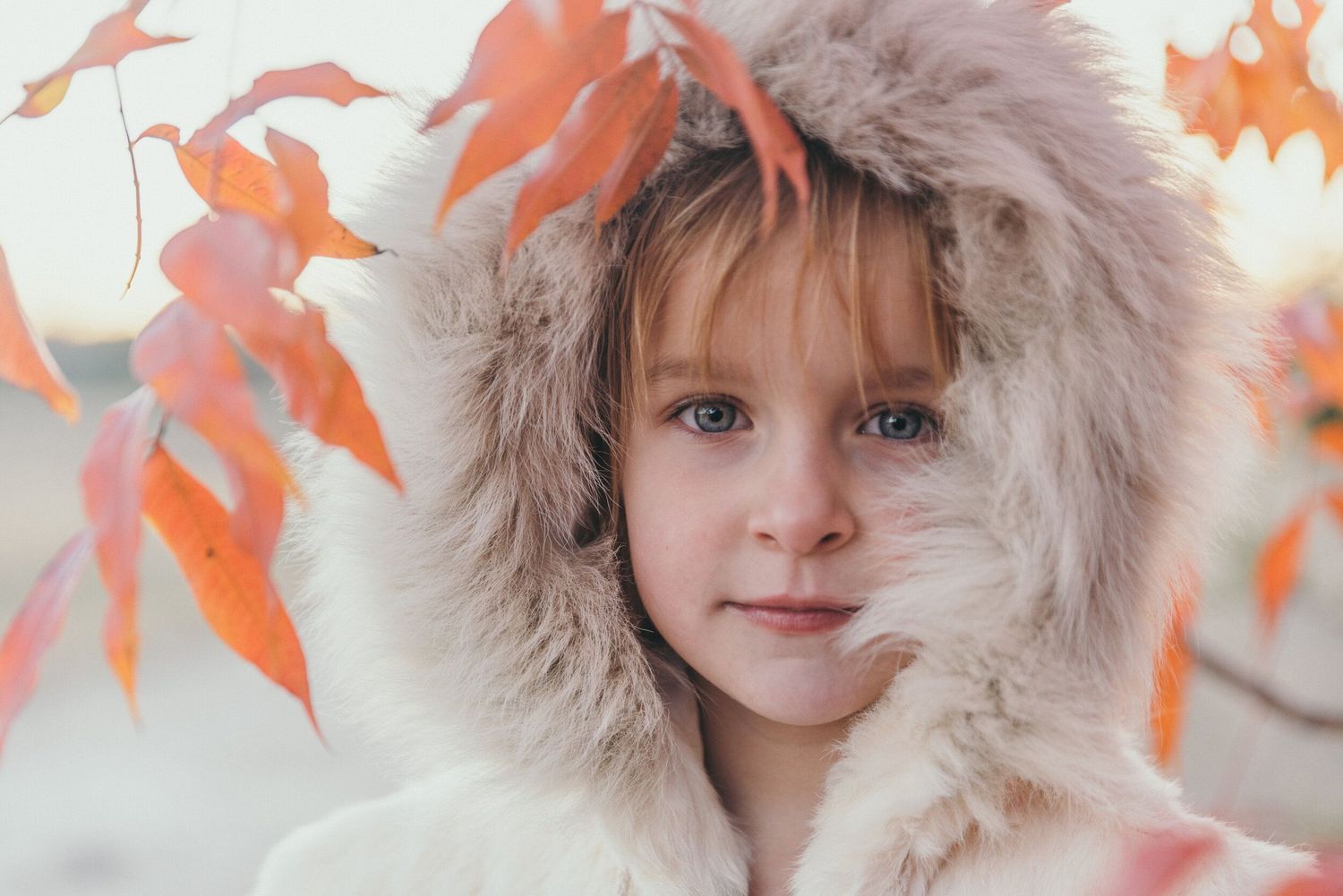 Close-up portrait of child wearing fur-trimmed hood with autumn leaves in background.