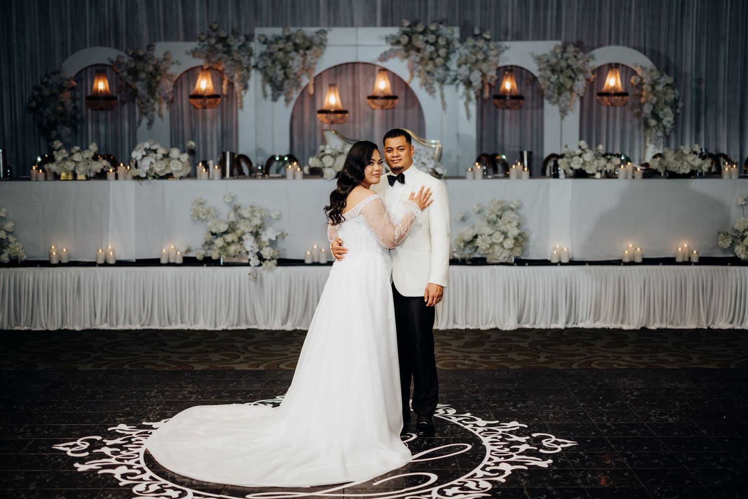 Elegant wedding couple poses on decorative floor pattern with illuminated backdrop and white drapery.