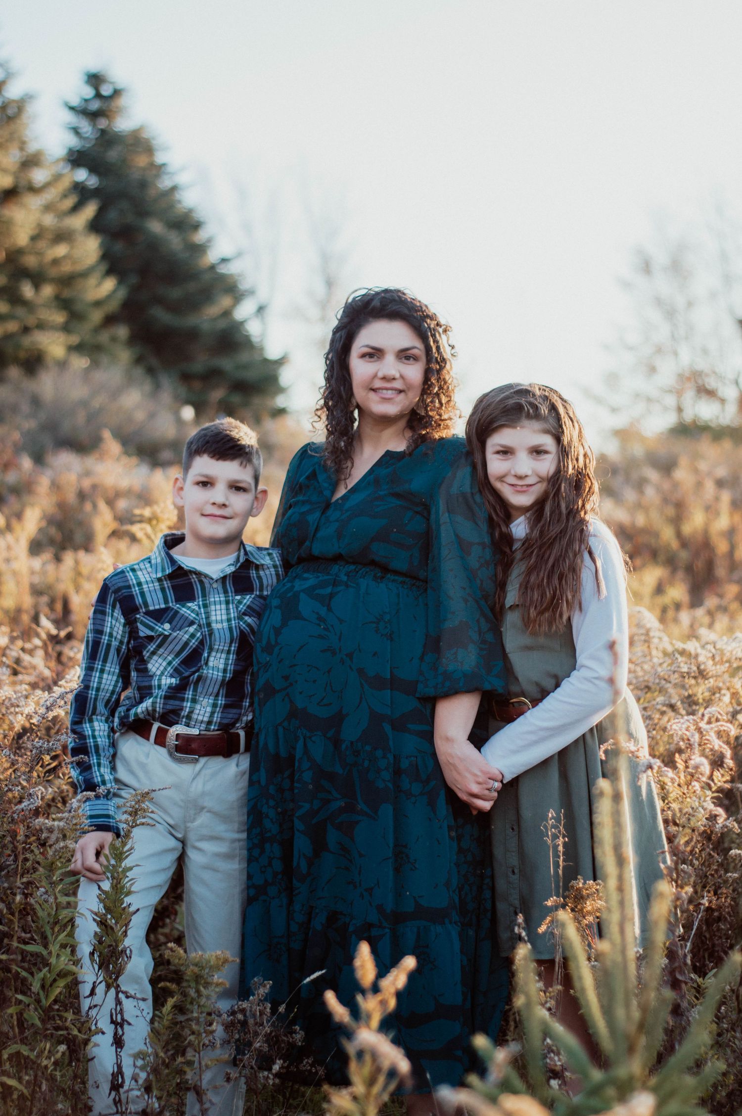 A pregnant mother and her two older children gaze confidently in a field of tall golden grass. Captured by M. Rose Photography.