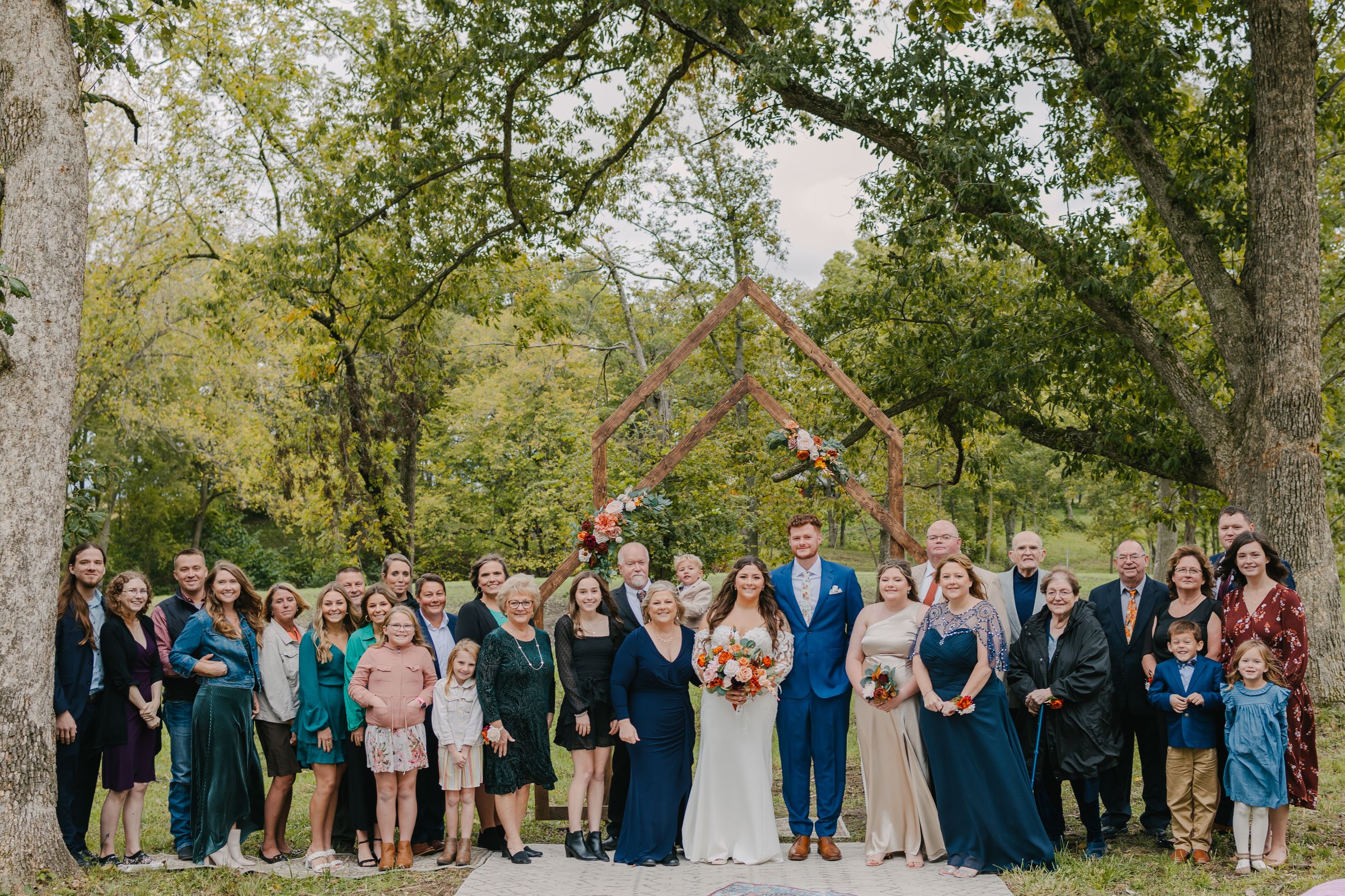 Large wedding party gathered outdoors for a formal group photo under tall trees.