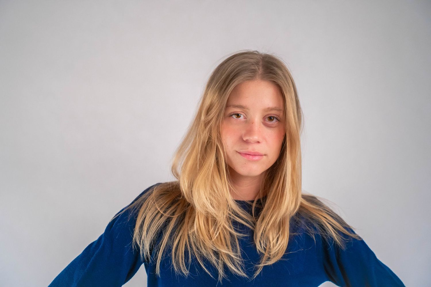 Young blonde girl with long hair wearing blue top against gray background.