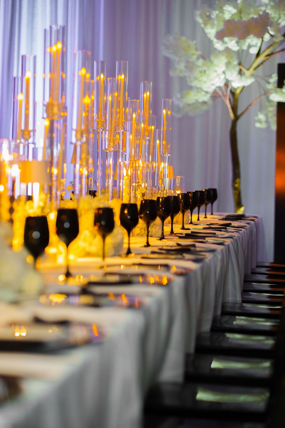 Elegant banquet table with black stemware, tall candle centerpieces, and white floral tree decor against draped backdrop.