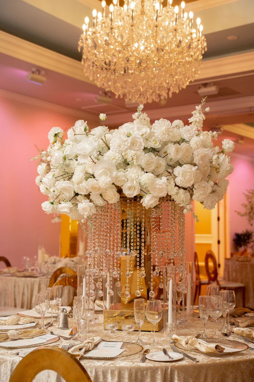 Elegant wedding reception centerpiece with white roses, crystal strands, gold vase, and chandelier in a pink-lit ballroom.