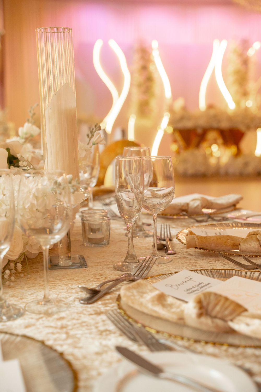 Elegant wedding reception table setting with gold accents, crystal glasses, white candles, and floral centerpieces in warm lighting.