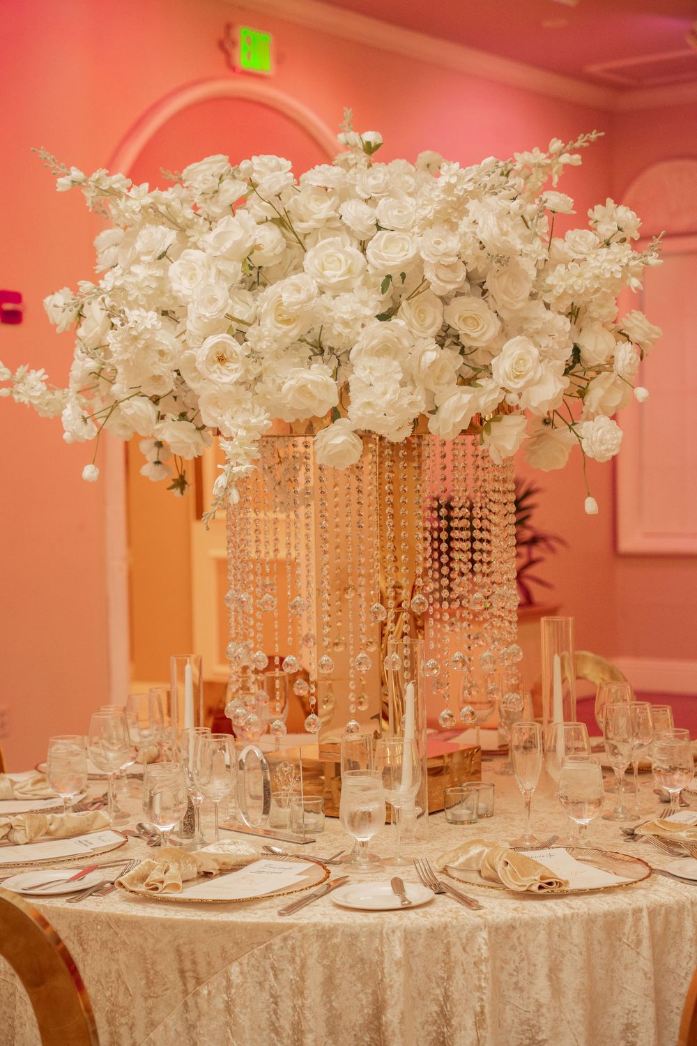 Elegant wedding reception table with tall crystal centerpiece adorned with white roses and candles on a gold lace tablecloth.