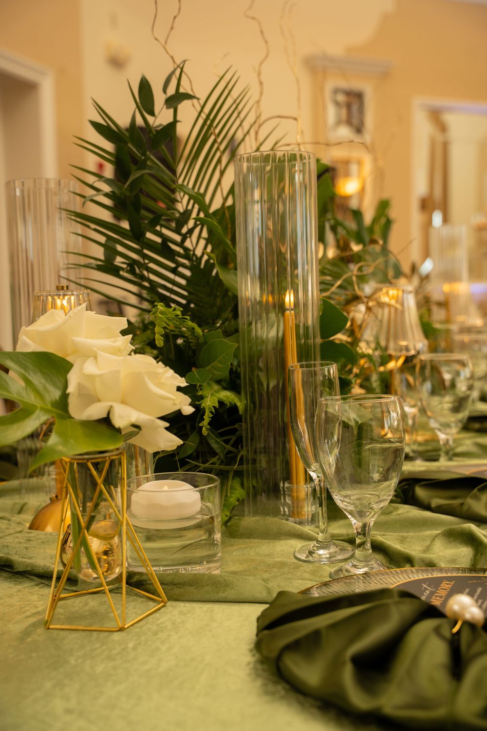 Elegant wedding table setting with green napkins, white roses, tall glass candles, and tropical greenery centerpiece.