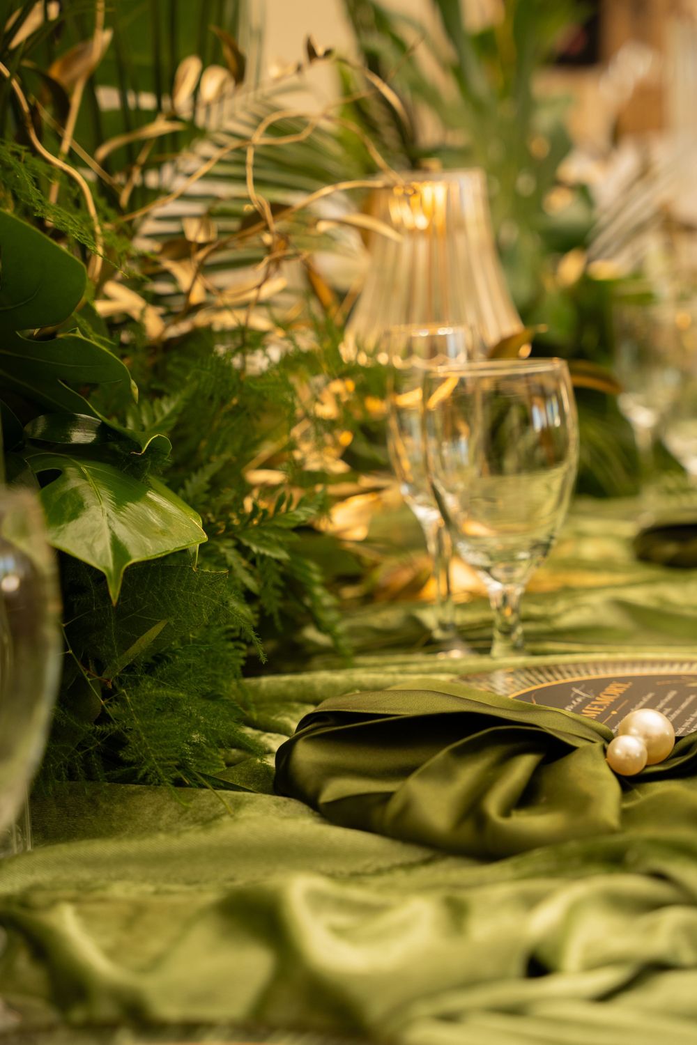 Elegant green and gold table setting with wine glasses, tropical leaves, and satin napkins for a luxury event.