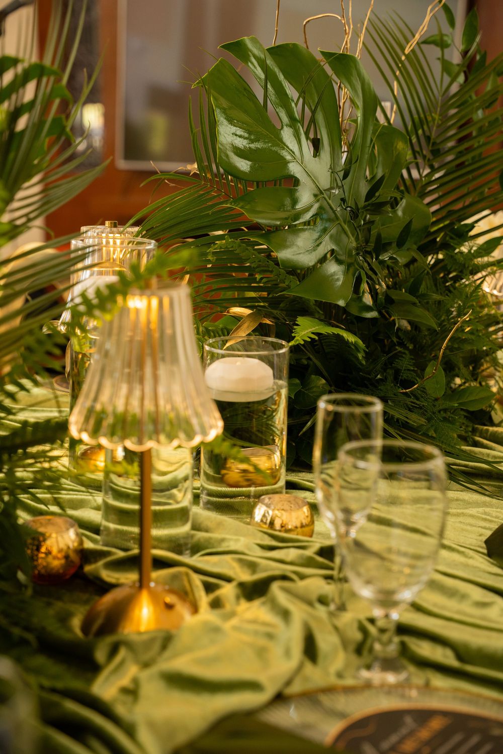 Elegant tropical table setting with monstera leaves, glowing gold lamp, floating candle, and green silk runner.