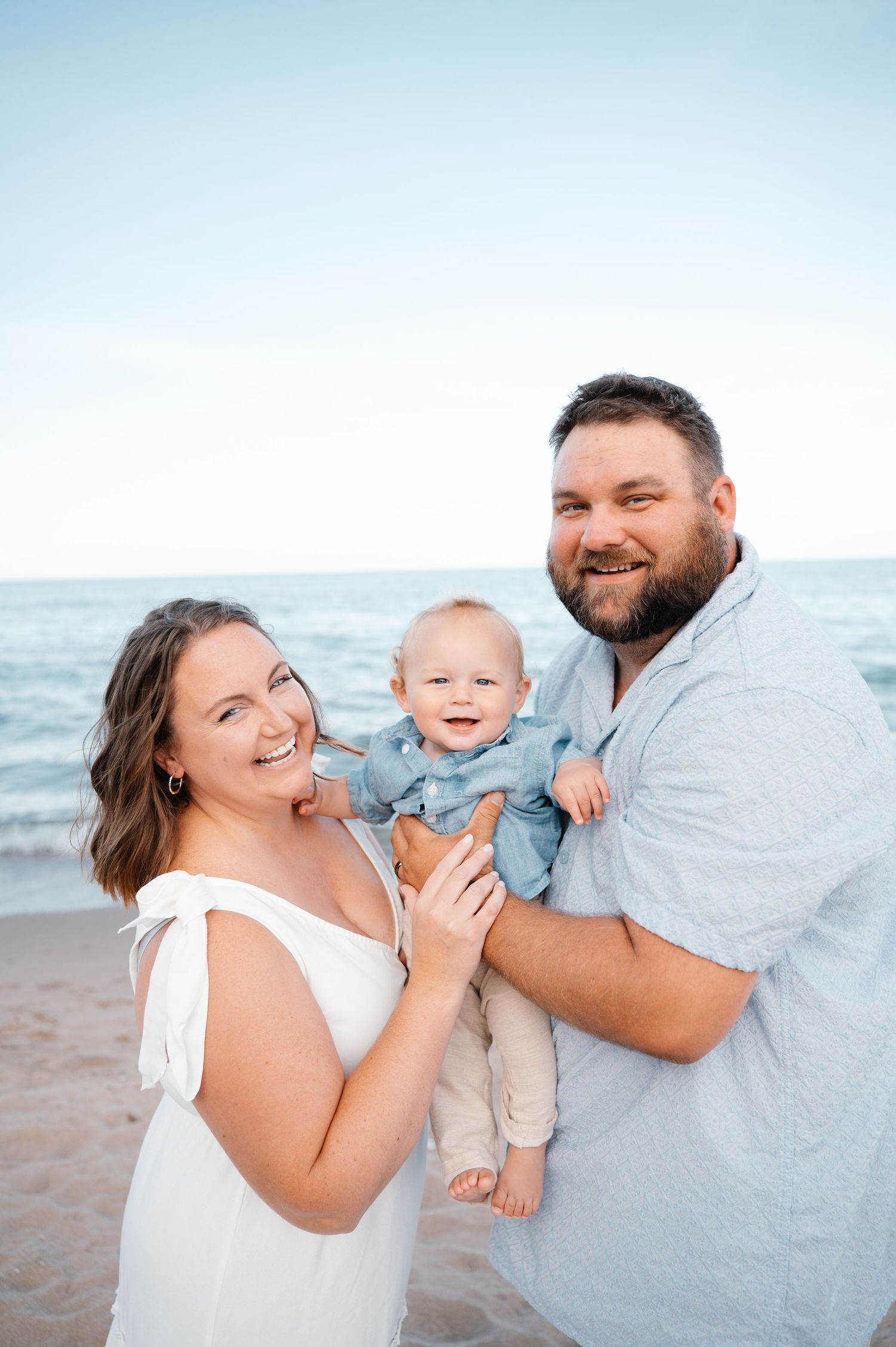 A smiling family of three with their happy baby poses together on a sandy beach at dusk.
