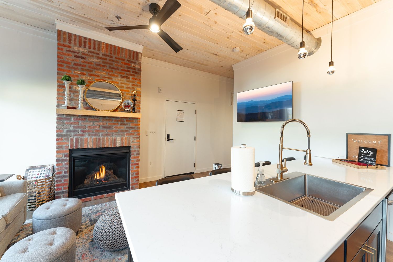 Modern kitchen interior with white countertop island, brick fireplace, and vaulted ceiling with exposed wood beams.