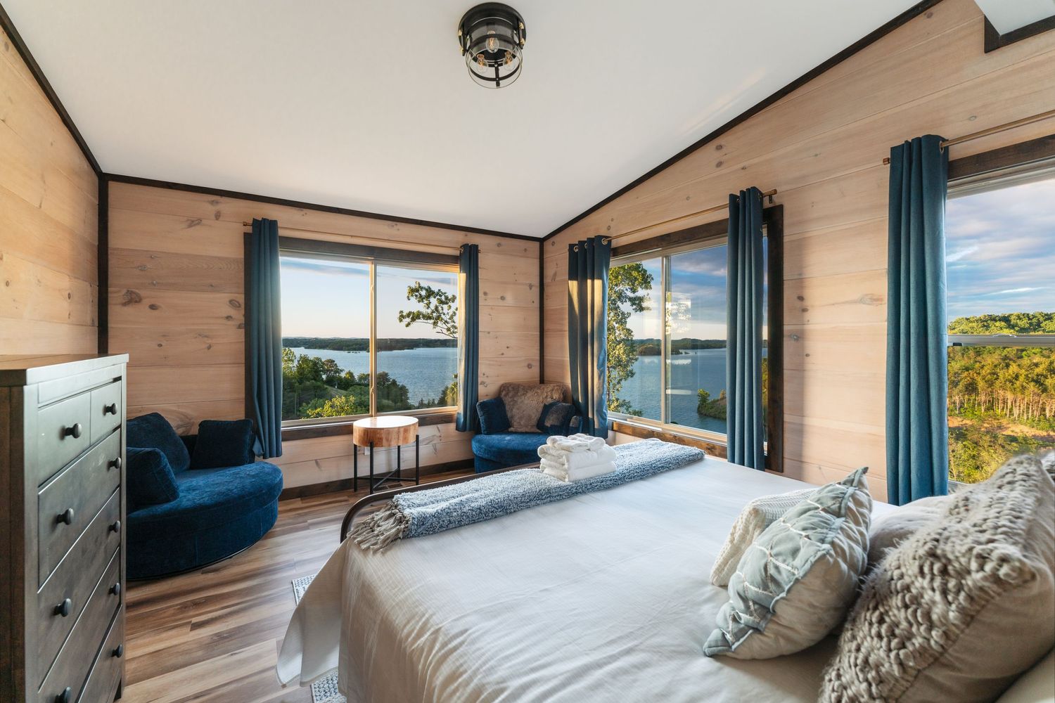 Modern bedroom with blue velvet chairs, wood walls, panoramic windows and ocean views features a white bed and teal curtains.