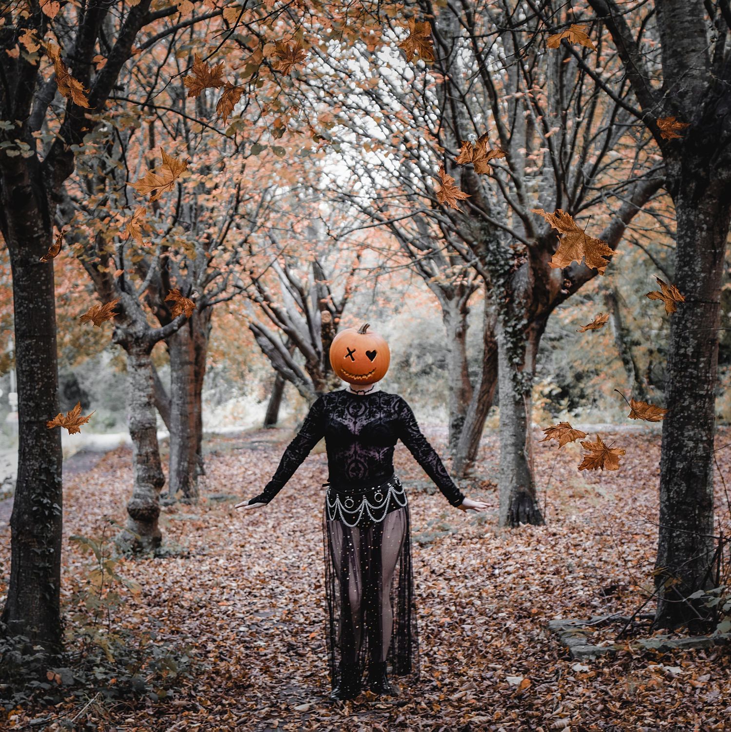 Eerie figure wearing black lace dress and jack-o-lantern head stands among fall trees with orange leaves.