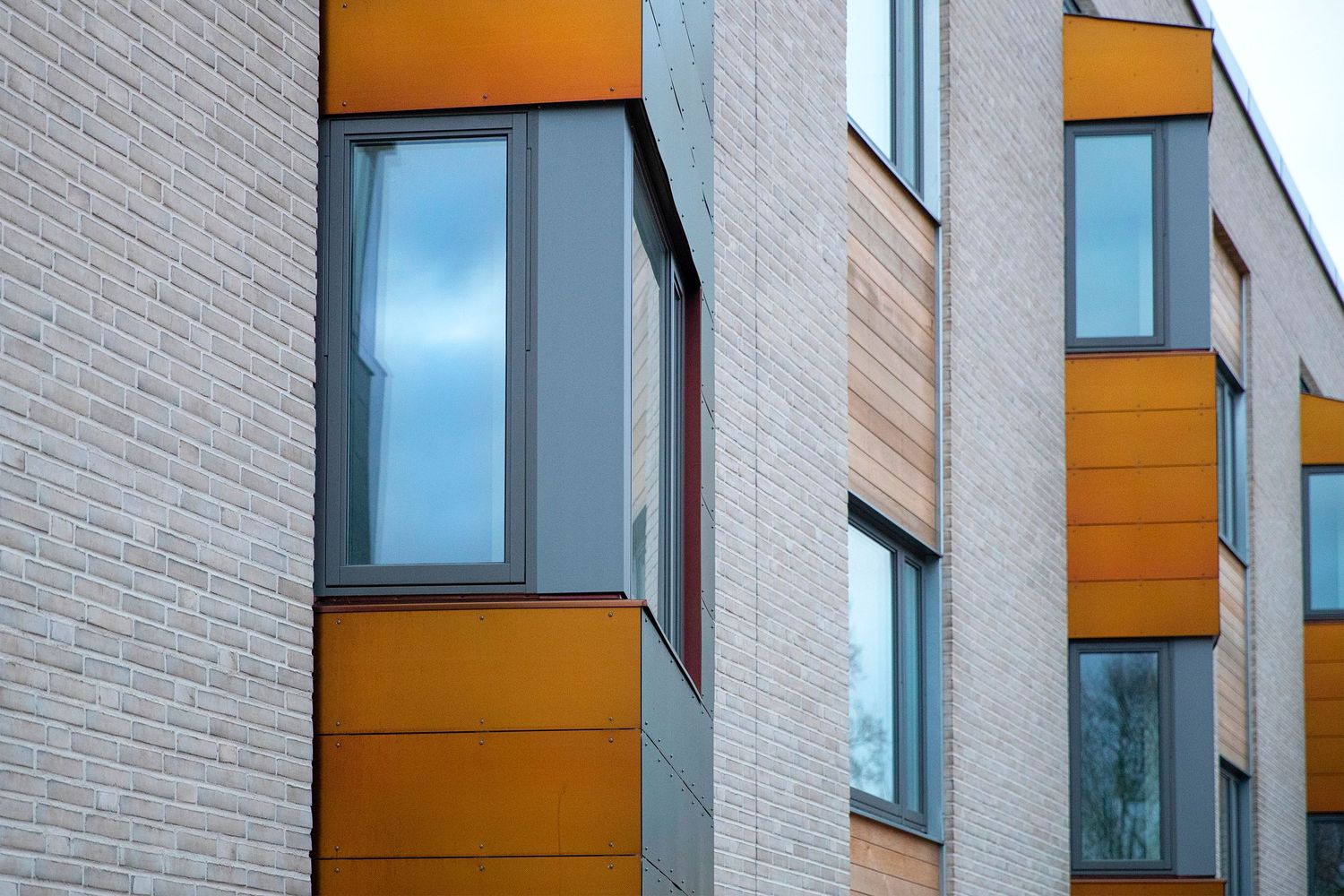 Modern apartment building exterior featuring orange accent panels and large windows with light gray brick facade.