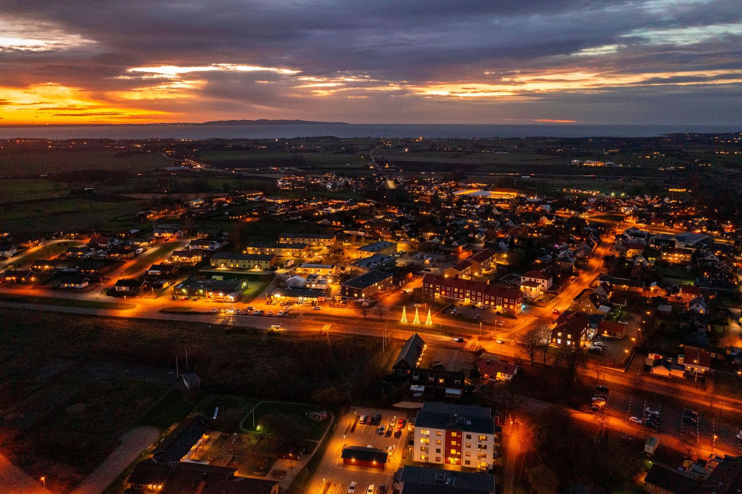 Aerial cityscape at dusk with glowing orange street lights illuminating neighborhoods under dramatic cloudy skies.