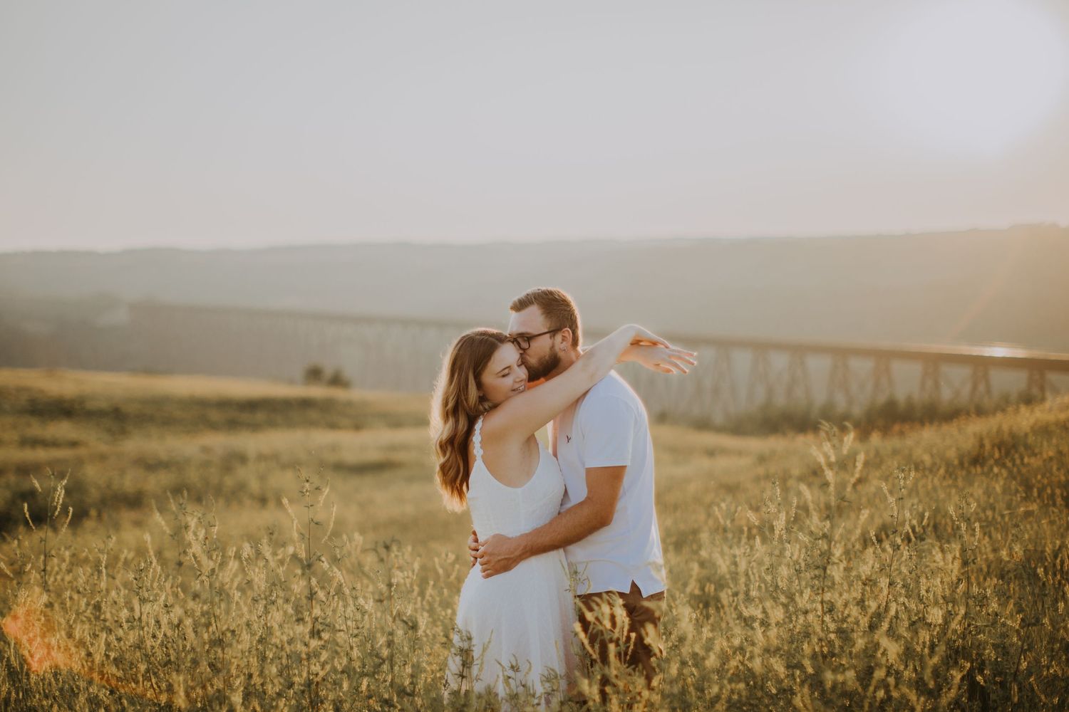A romantic couple embraces in a sun-lit field with a bridge visible in the distance.
