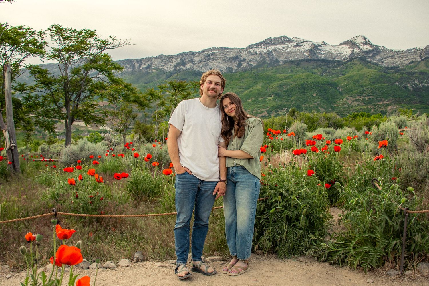 Girl lays head on boys shoulder in a field of poppies with an Utah mountain landscape in the background.