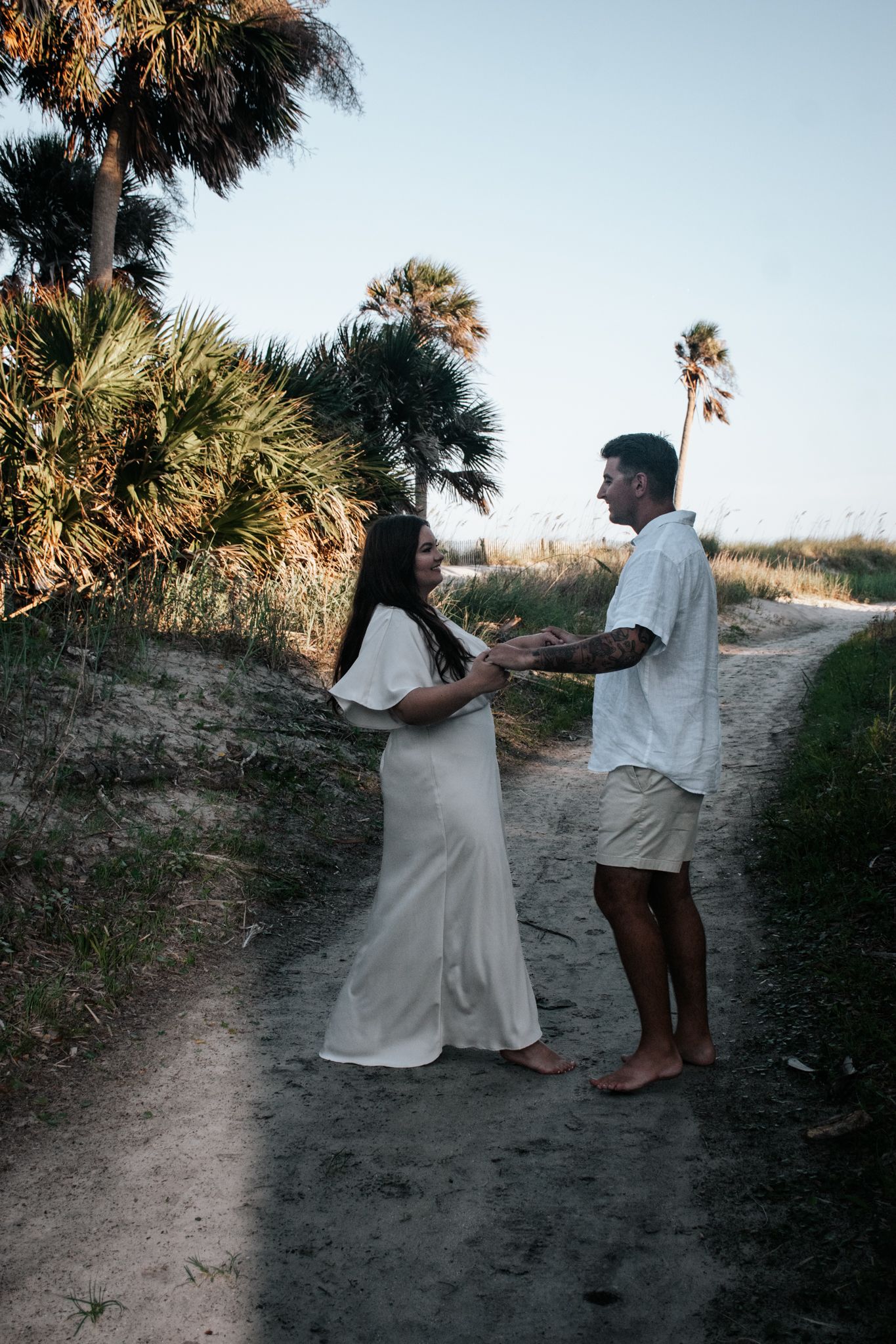 A couple holding hands on a sandy path surrounded by palm trees during a photoshoot.