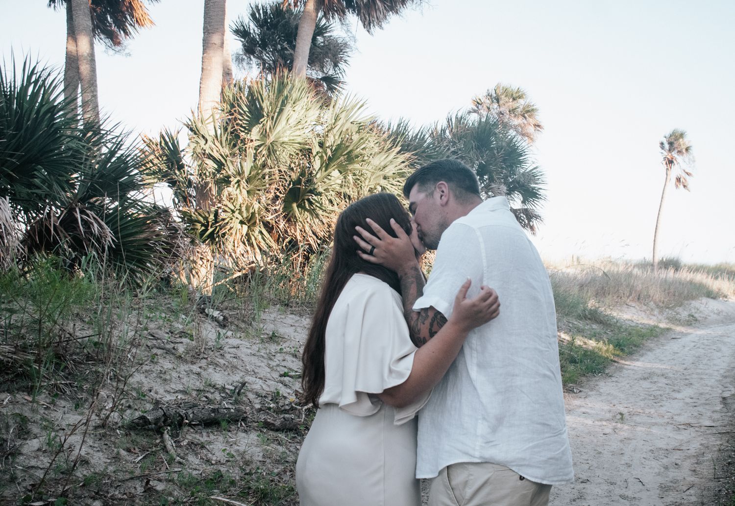 A couple sharing an intimate embrace on a coastal path with palm trees.