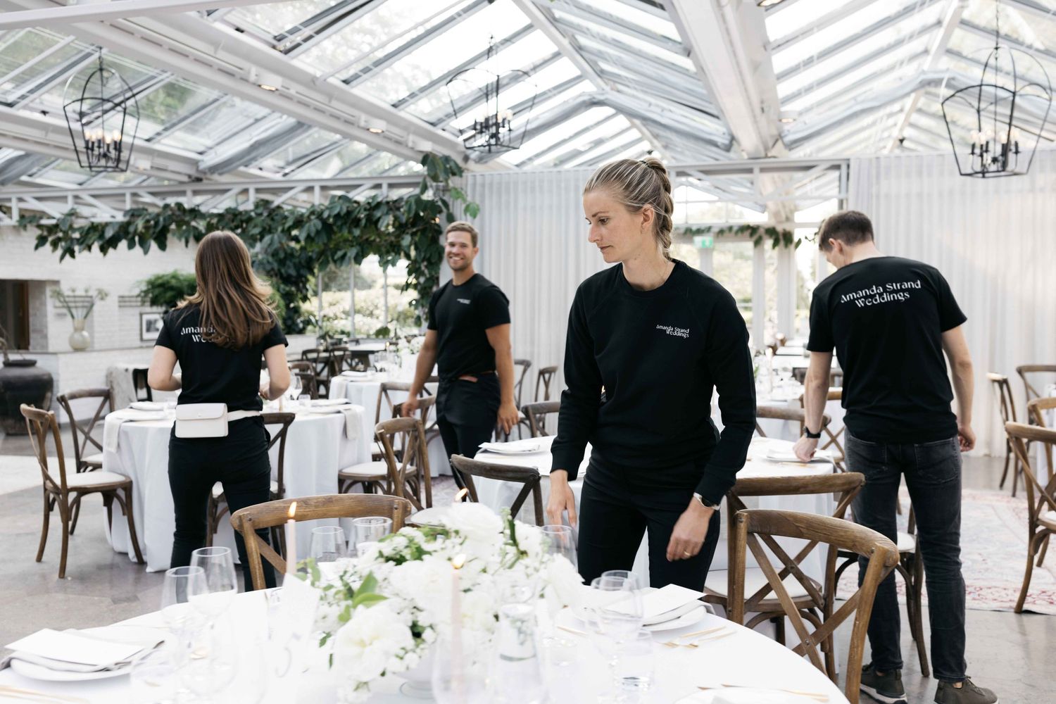Staff in black uniforms prepare elegant table settings in a bright conservatory venue.