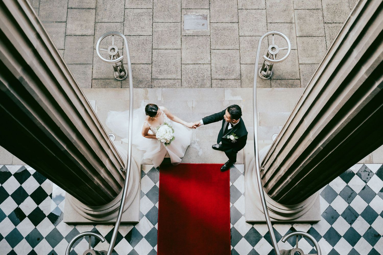 Aerial view of couple on red carpet between classic architectural columns with checkered floor.