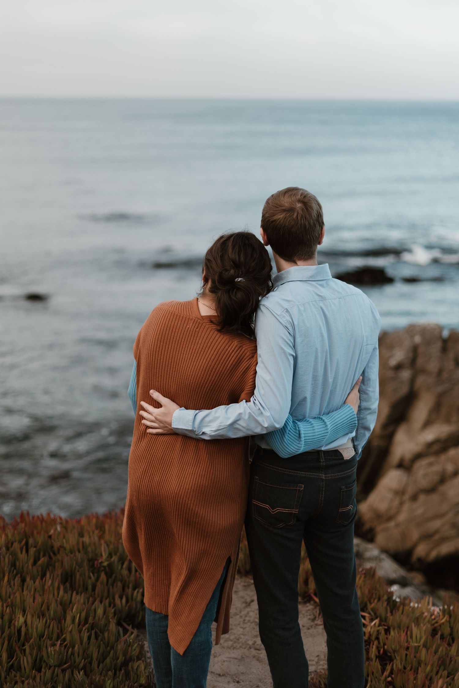 Couple embracing while looking out at the ocean from a rocky coastal viewpoint.
