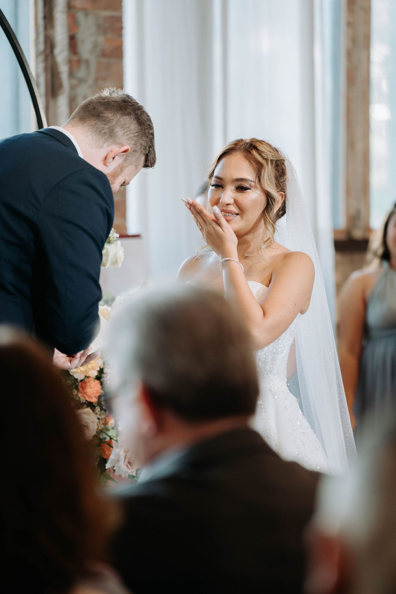 Bride shows emotional reaction during wedding ceremony in light-filled venue.