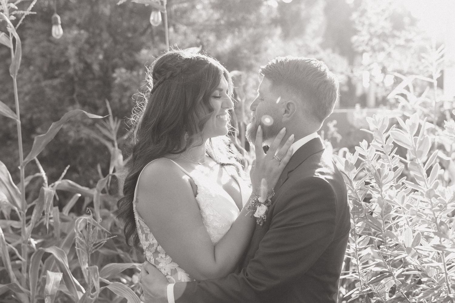 Couple in wedding attire standing close outdoors, facing each other and smiling amid greenery in soft natural light.