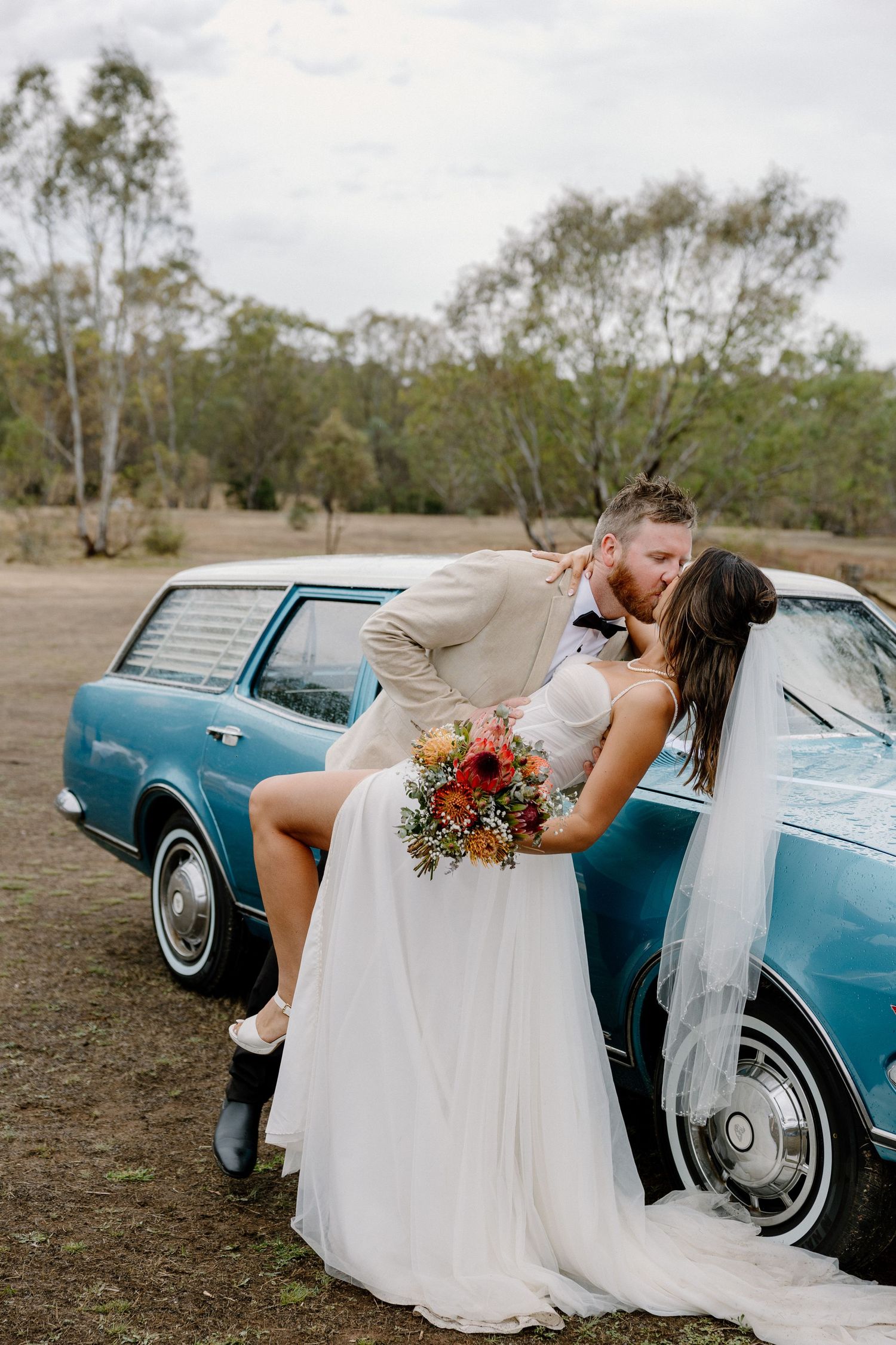 Newlywed couple sharing romantic moment against vintage blue car in rustic Bendigo setting.