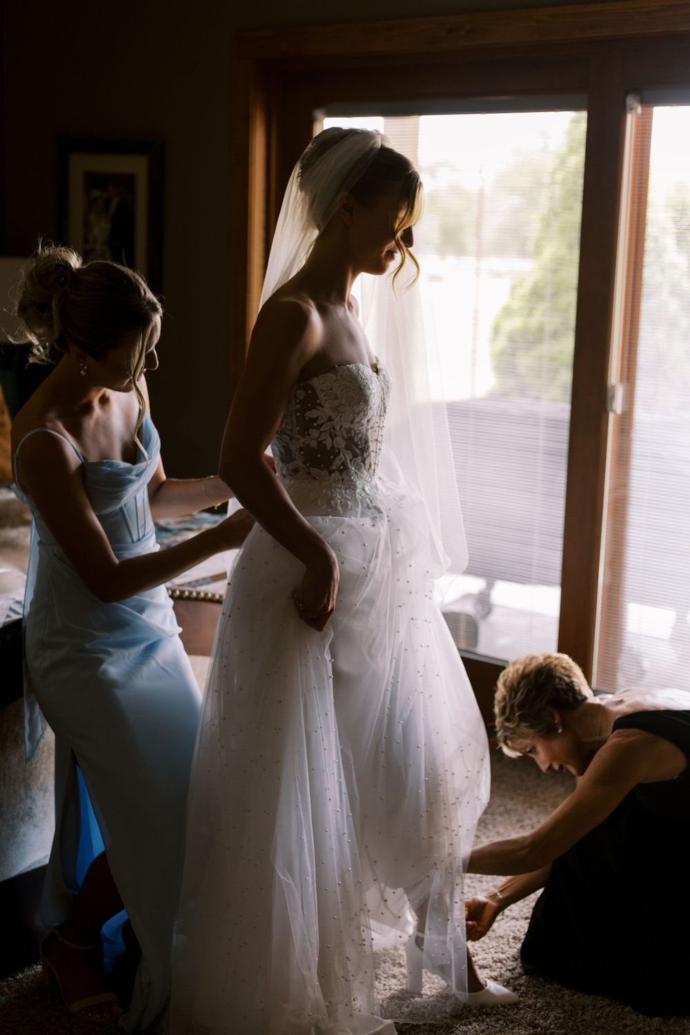 Wedding dress fitting moment as attendants help with final adjustments in sunlit room.