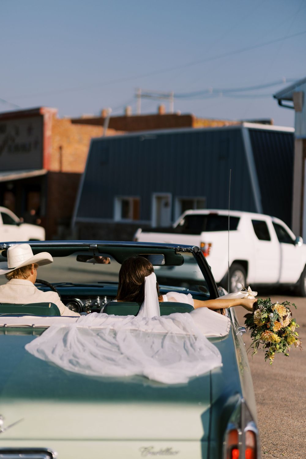 Wedding dress flowing from classic car with floral bouquet.