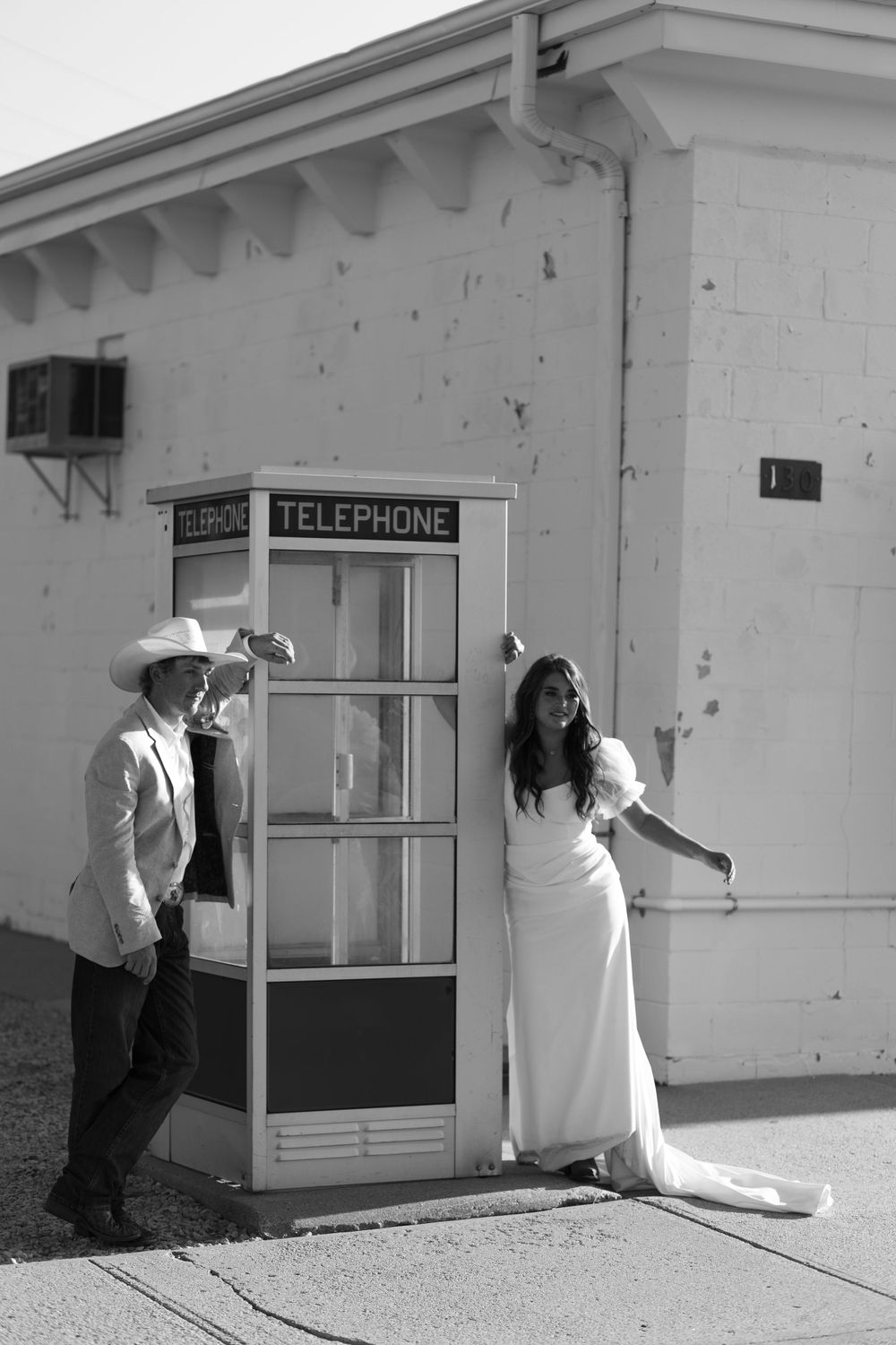 A couple poses playfully near a vintage telephone booth in black and white.