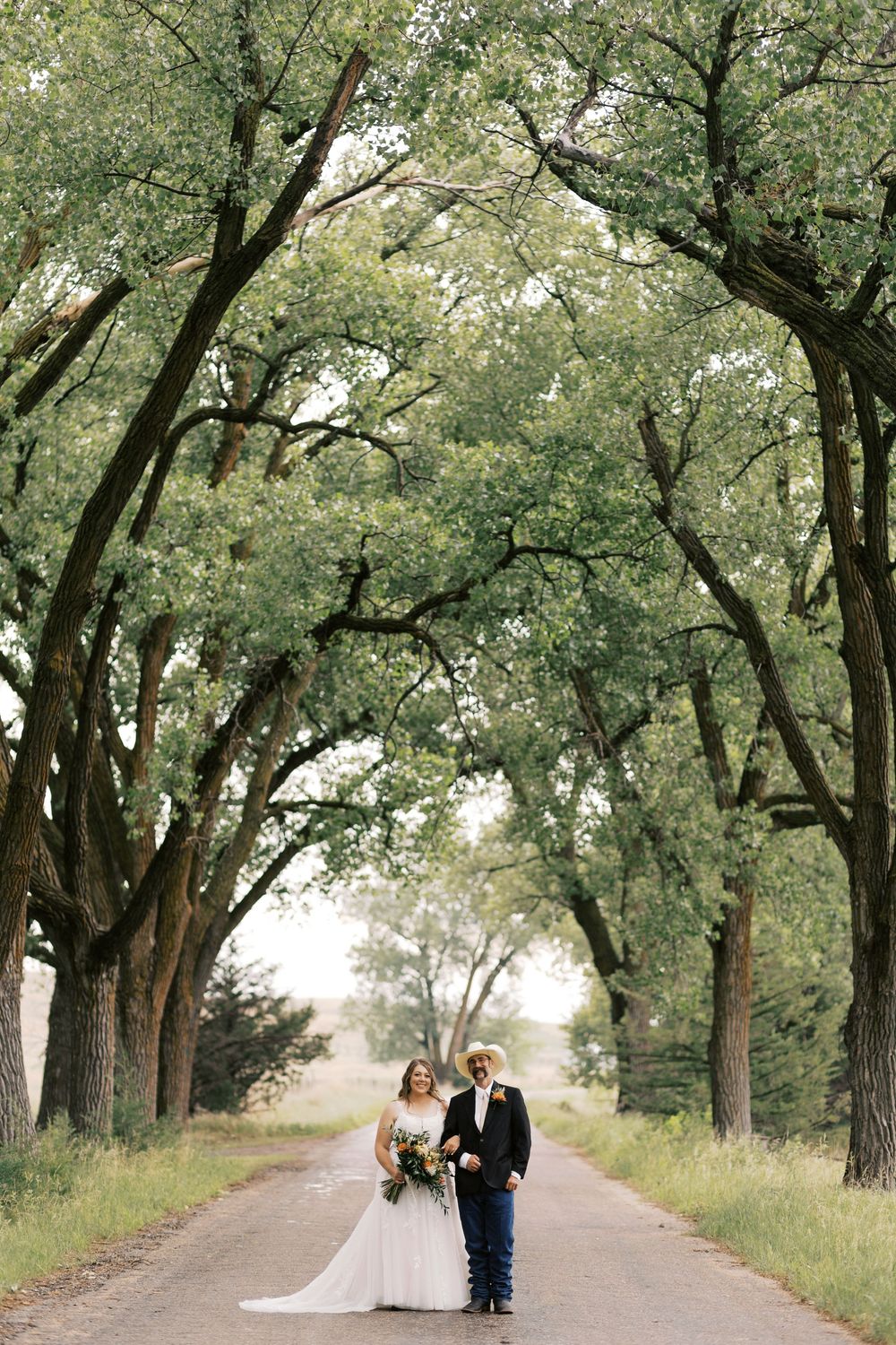 Bride and groom stand on a tree-lined road surrounded by lush green oak trees on their wedding day.