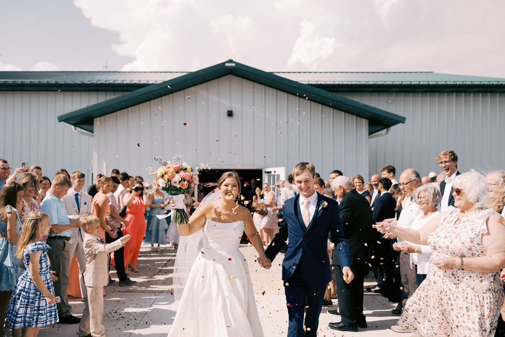 Joyful bride and groom exit white barn wedding venue as guests celebrate with confetti toss.