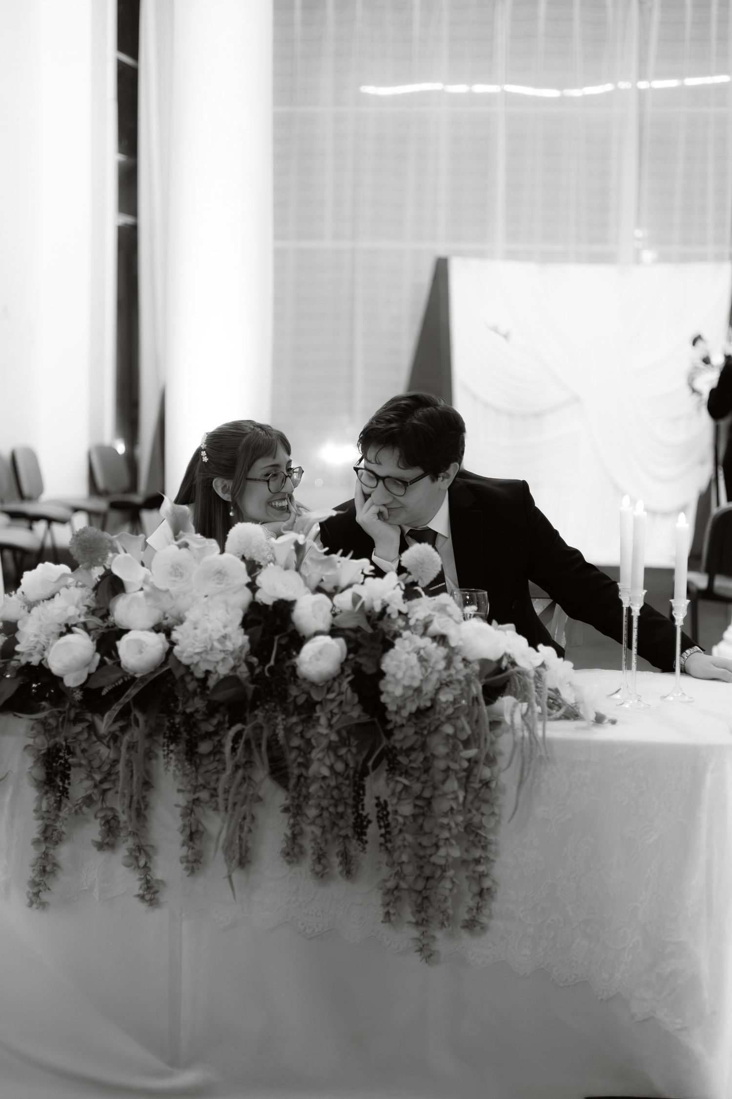 Black and white photo of newlywed couple laughing at floral-decorated sweetheart table at reception.