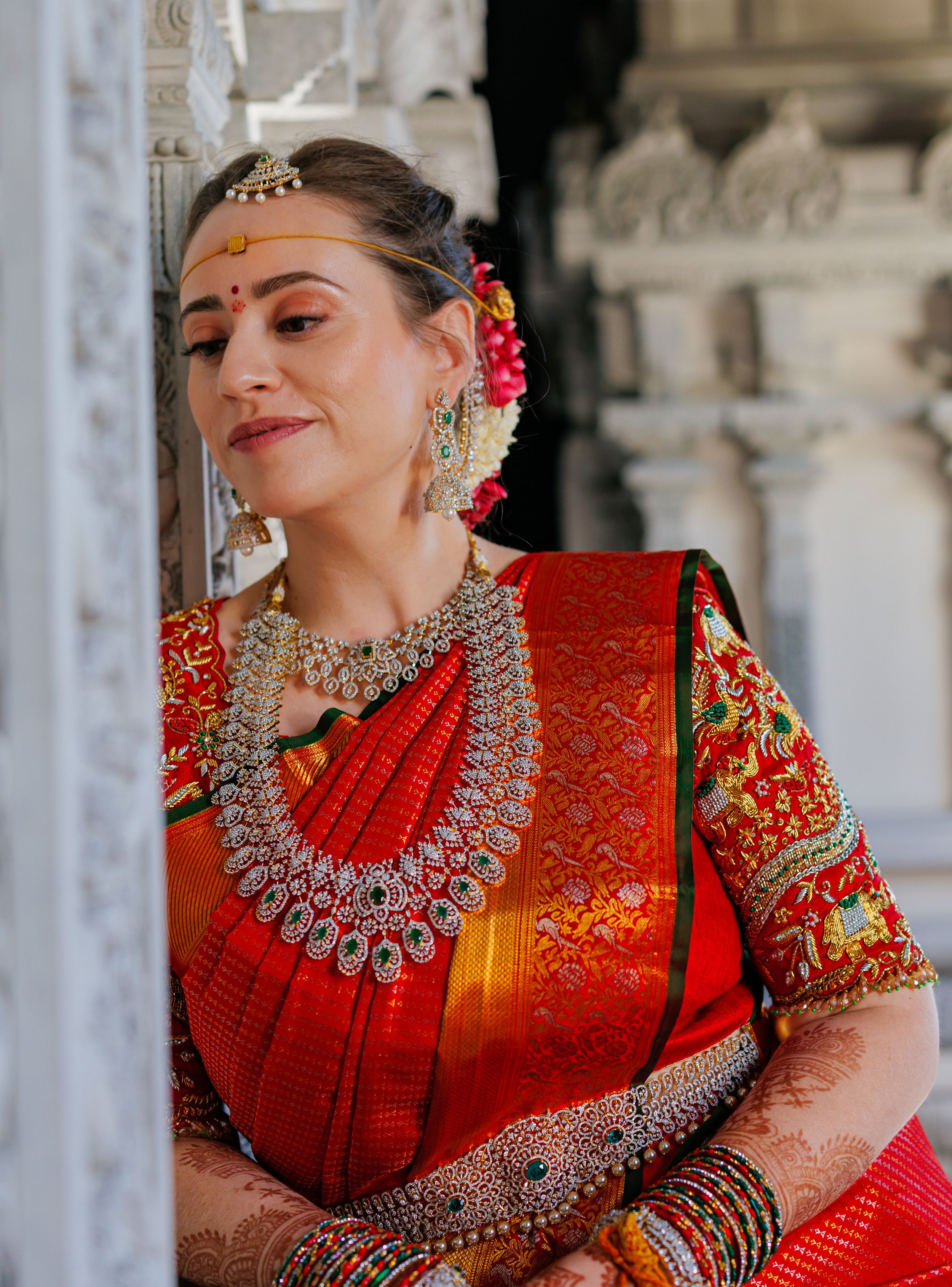 A bride in a red silk saree with gold jewelry, maang tikka, and floral hair accessories poses at a temple.