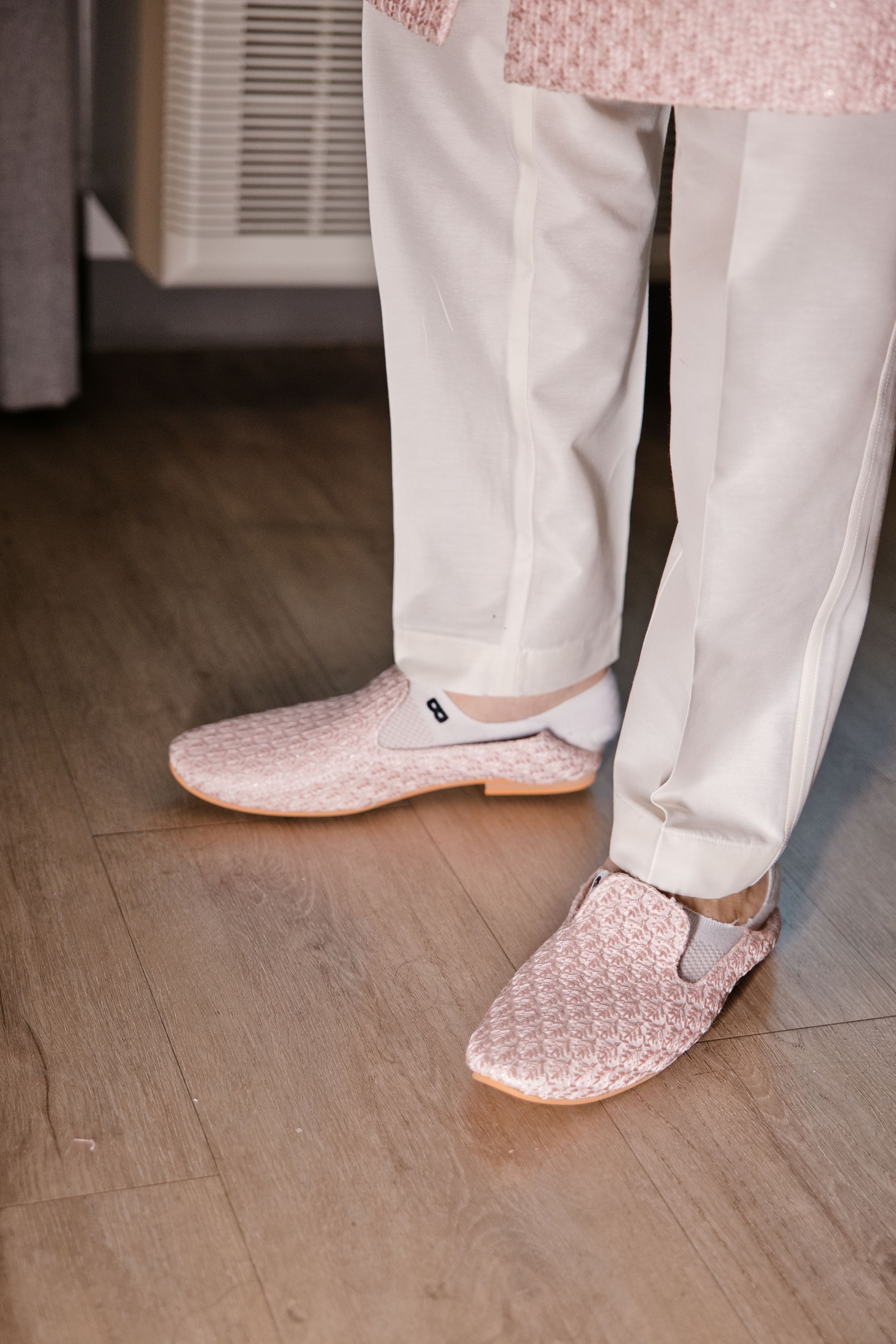 Close-up of a groom wearing white trousers and pink woven loafers on a wooden floor.