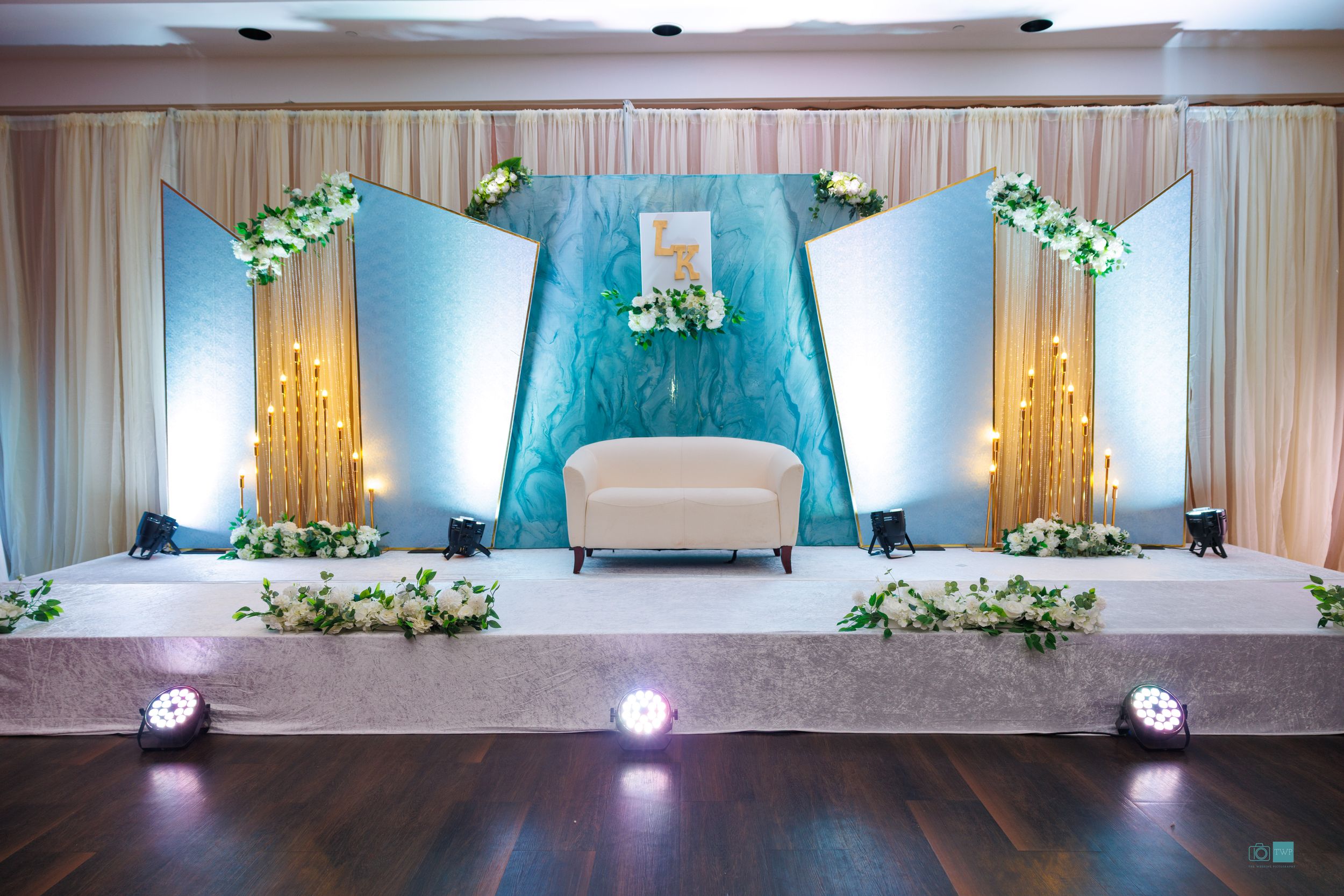 Elegant wedding stage with teal backdrop, white sofa, floral garlands, and golden candelabras in a decorated banquet hall.