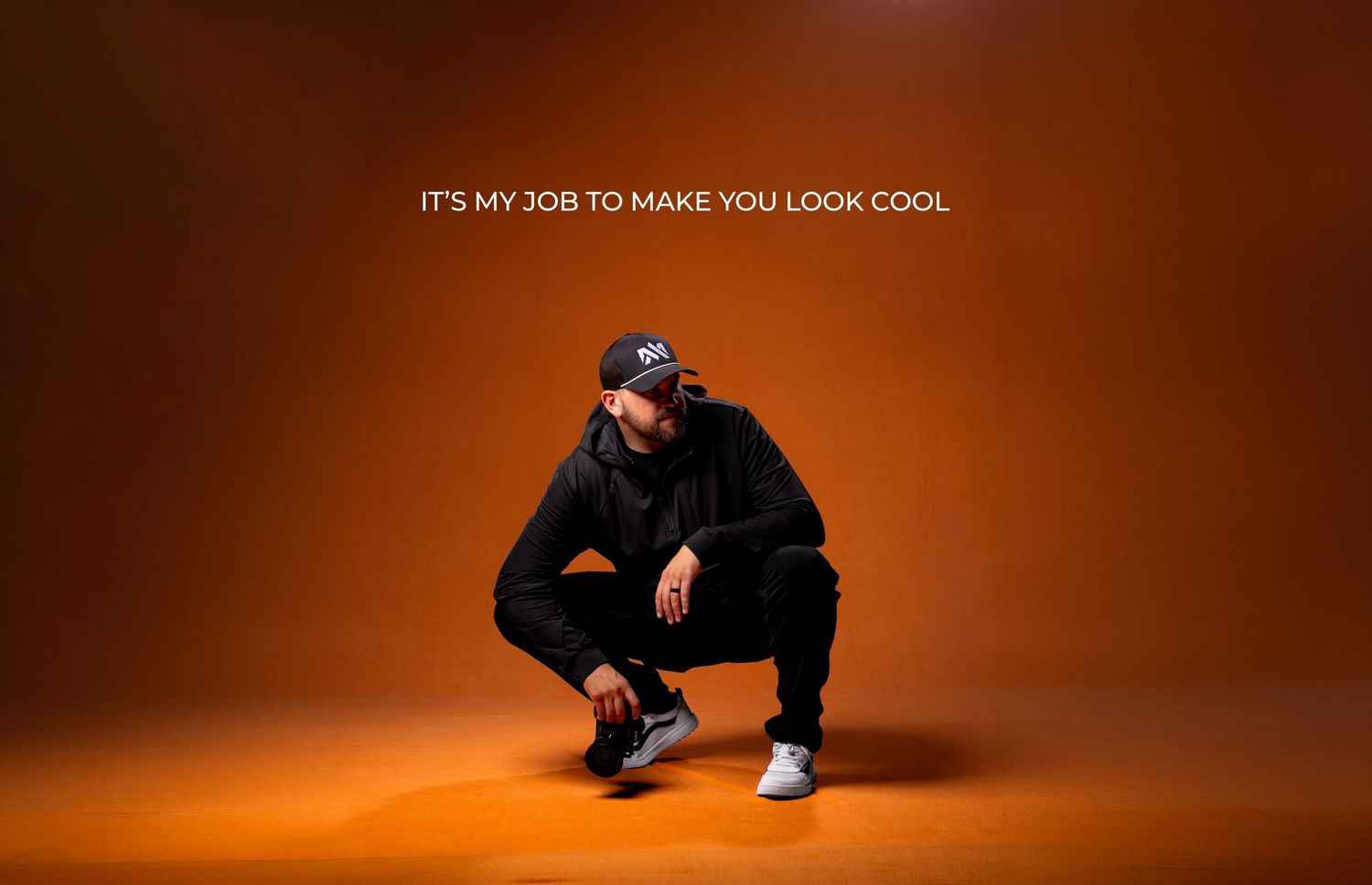 A confident man crouching in all-black outfit against orange background with text Its My Job To Make You Look Cool.