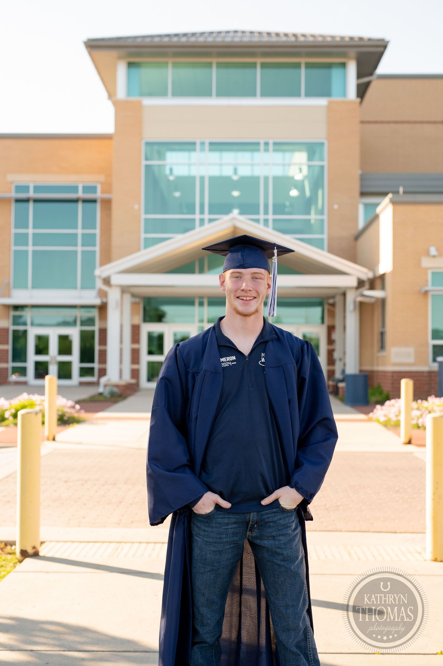 Graduate in navy graduation attire stands with hands in pockets outside modern building entrance.