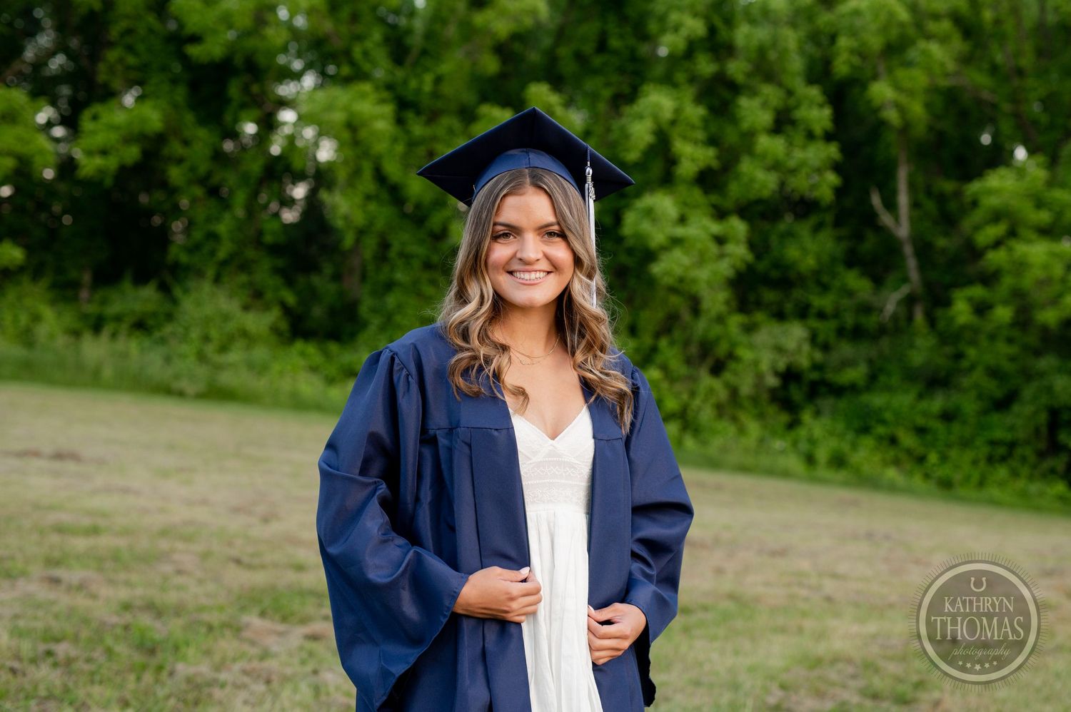 Graduate in navy blue cap and gown stands against green foliage background.