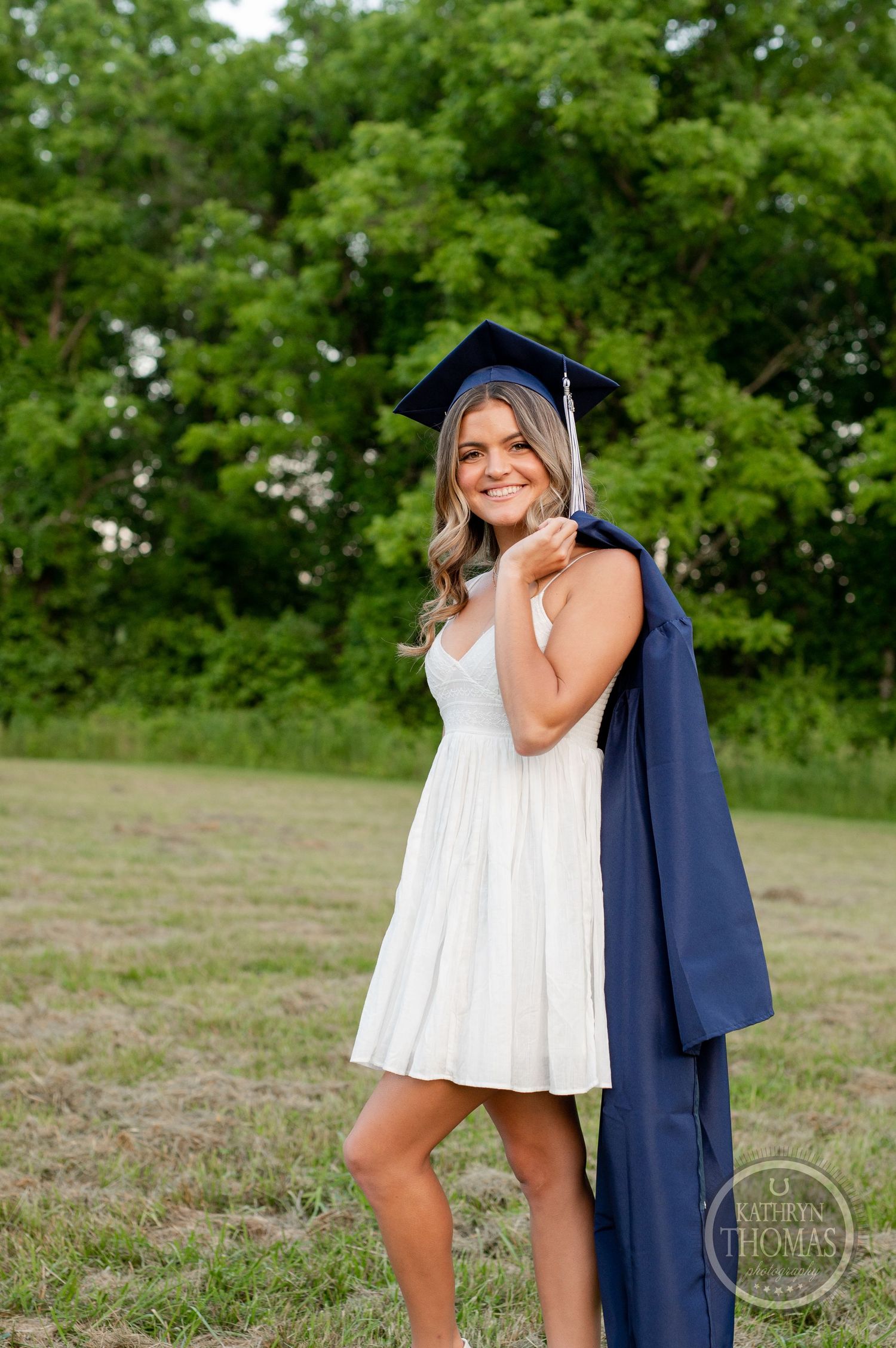 Graduate in white dress and navy cap poses playfully with gown in outdoor setting.