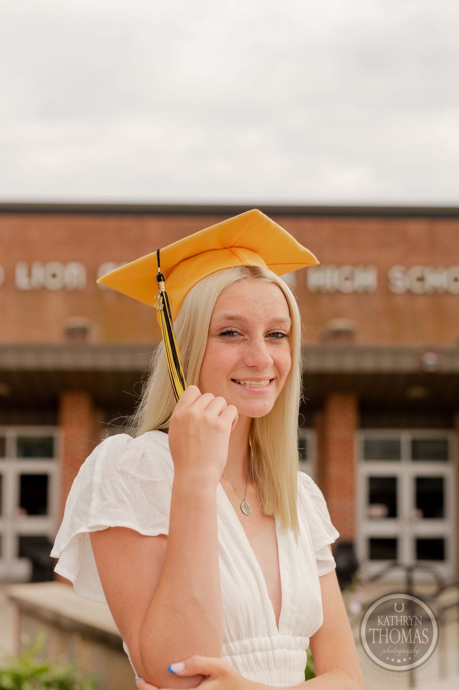 Graduate wearing gold cap with tassel poses outside brick school building.