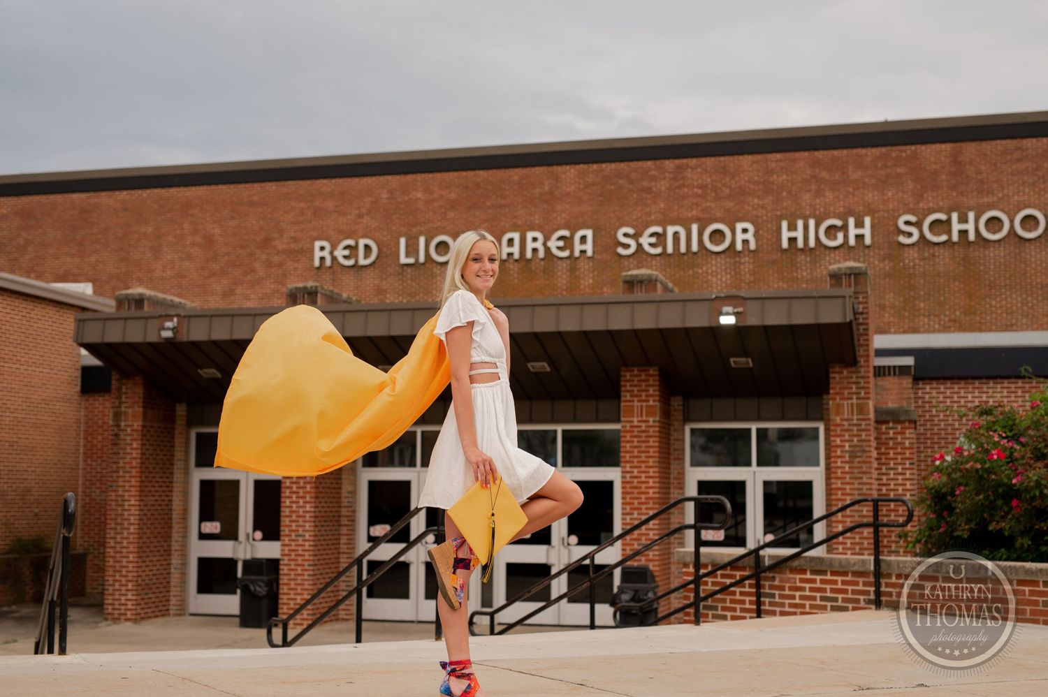 Graduate in flowing yellow graduation gown twirls in front of brick school building.