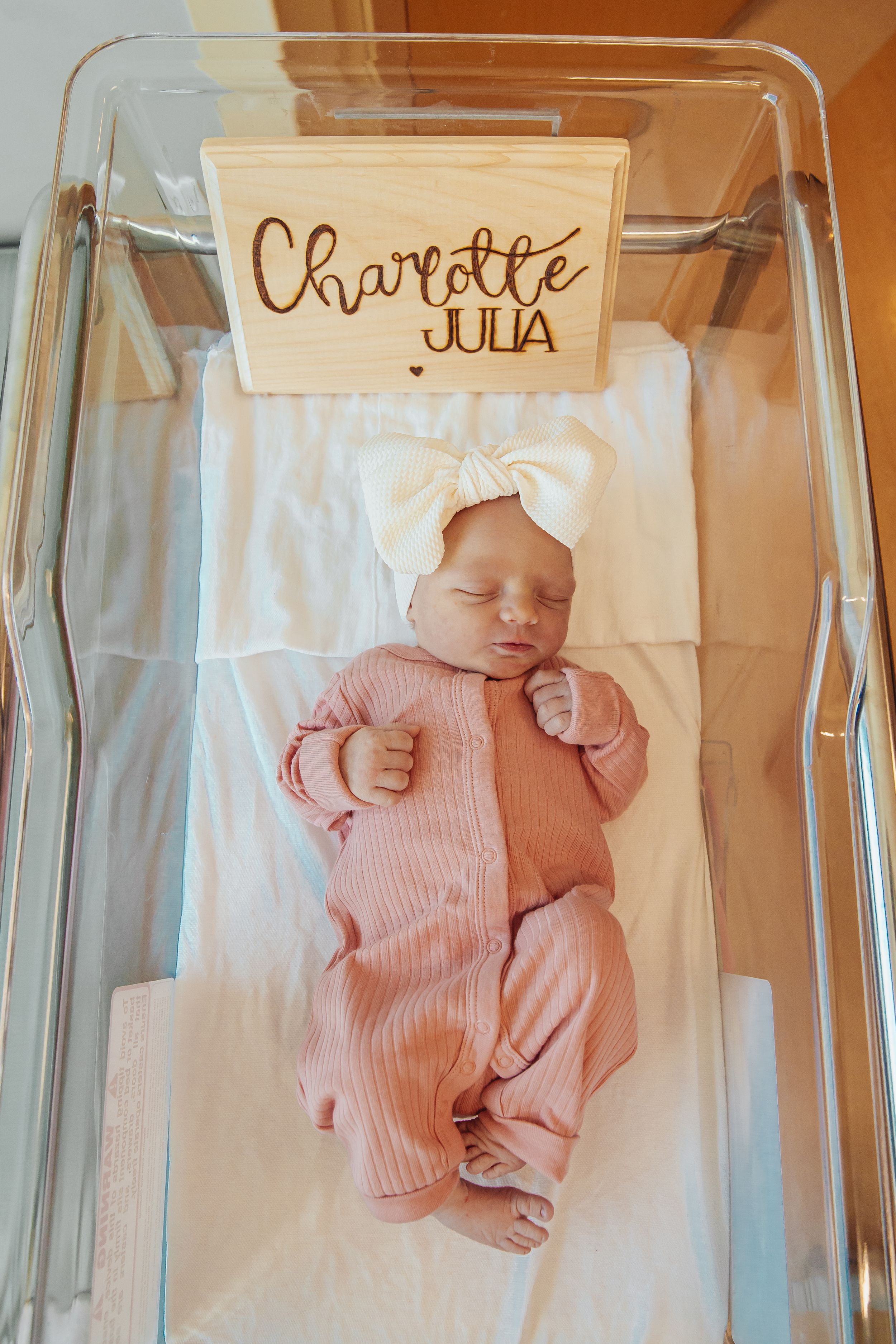 Newborn baby wearing pink sleepwear rests peacefully in a hospital bassinet.