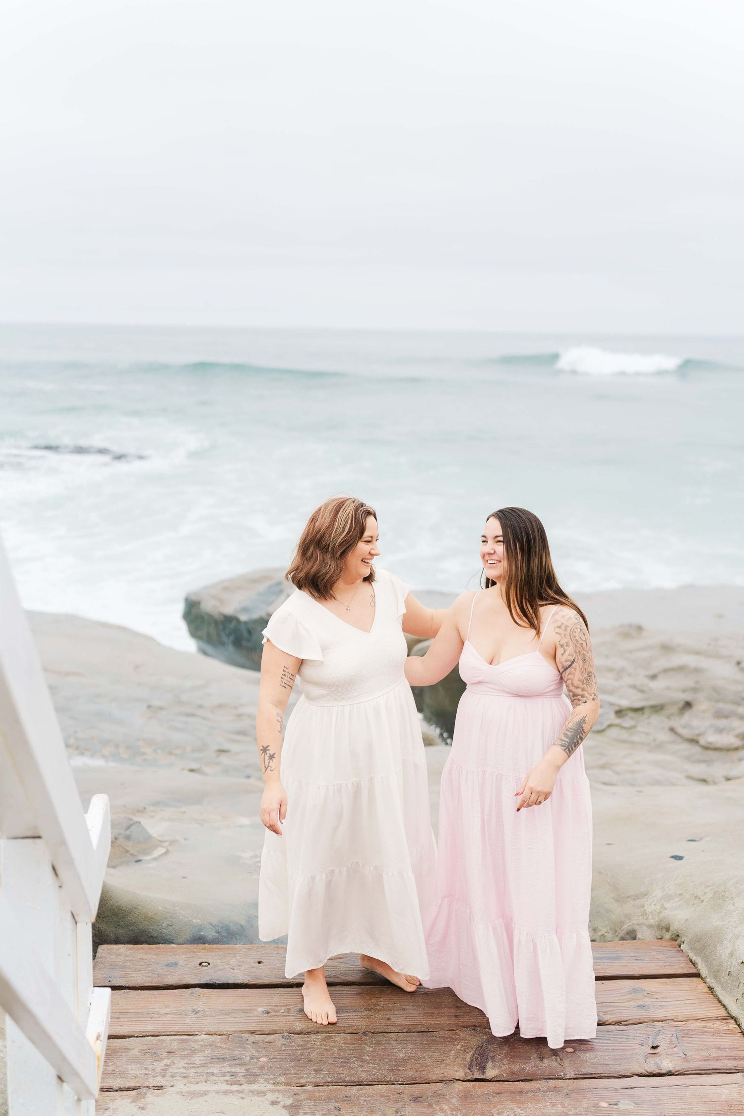 Two people in flowing pastel dresses share a joyful moment while standing on wooden steps by the ocean.