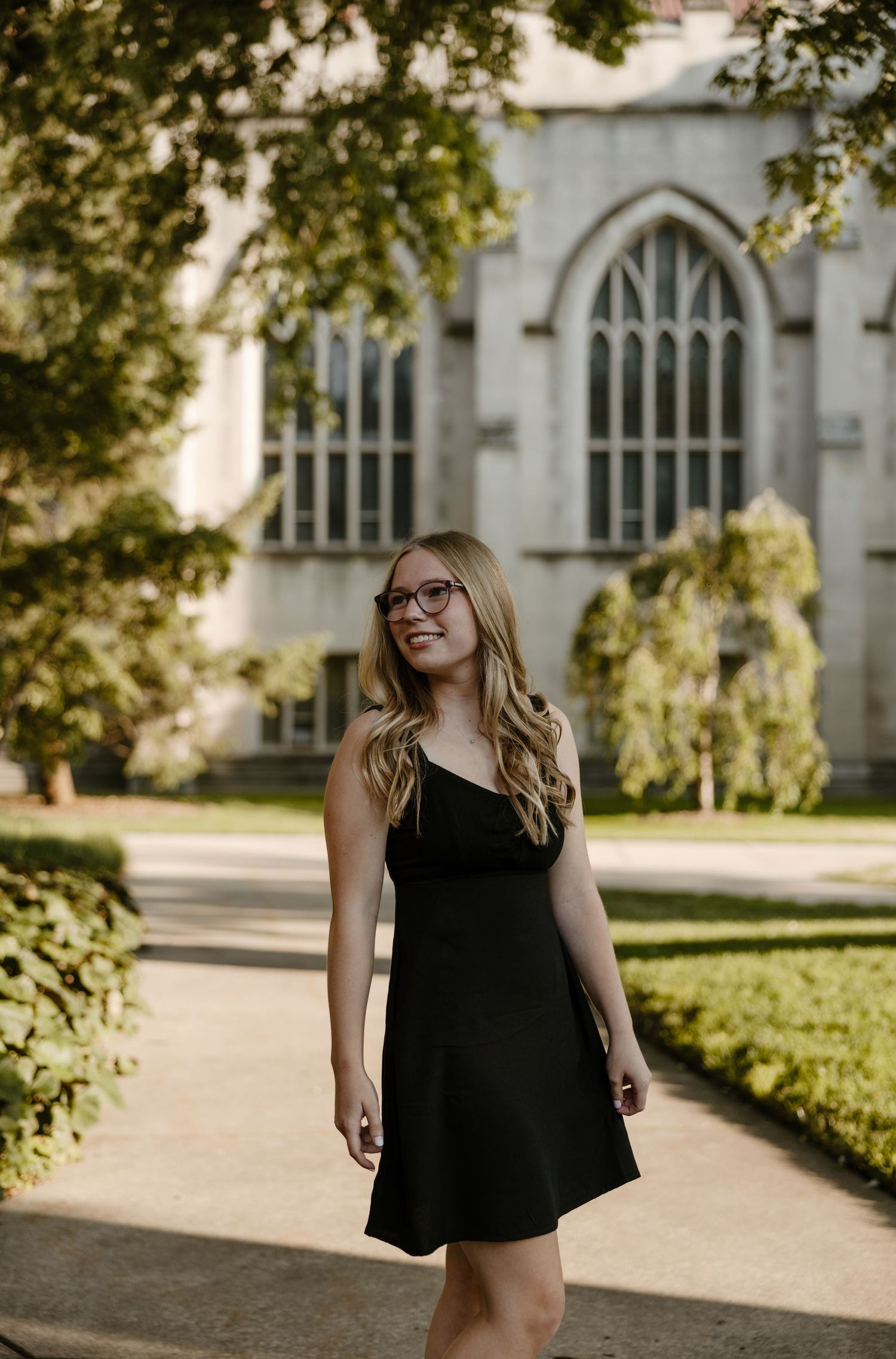 A young woman with long blonde hair wearing a black dress stands in front of a stone building with arched windows. Fort Worth Texas.