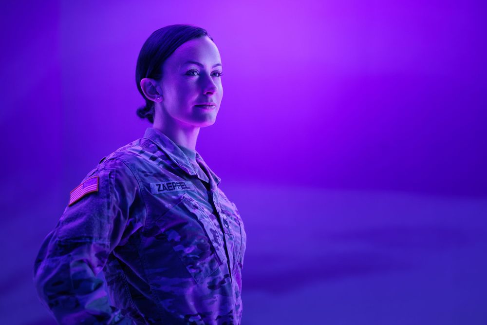 Service member in military uniform stands proudly against purple backdrop.