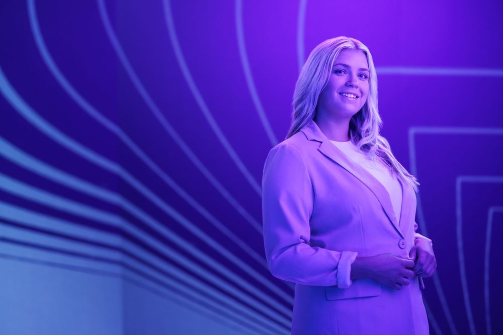Professional in light purple blazer stands against curved light pattern background.