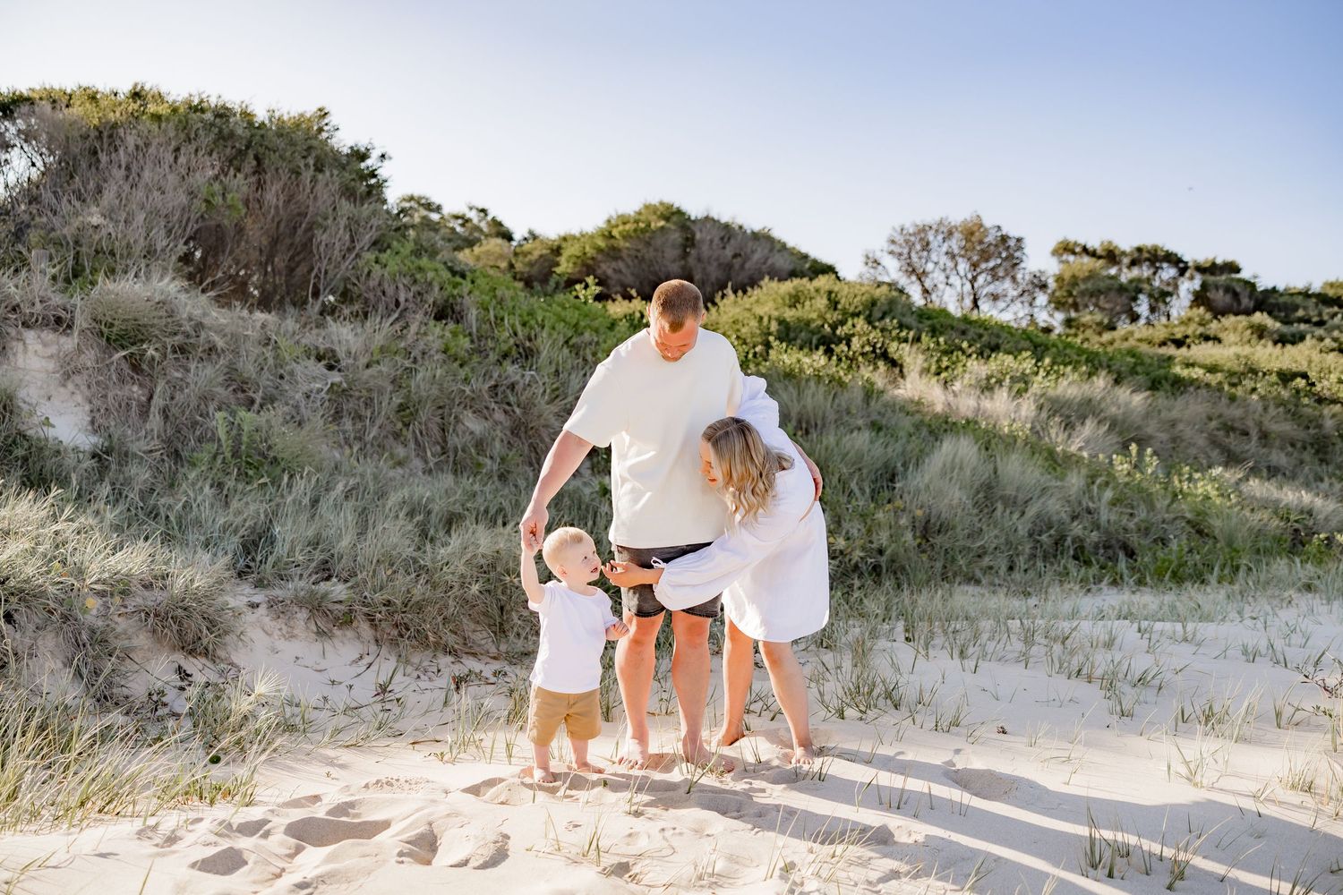 Family wearing coordinated white outfits playing on sandy dunes at beach.