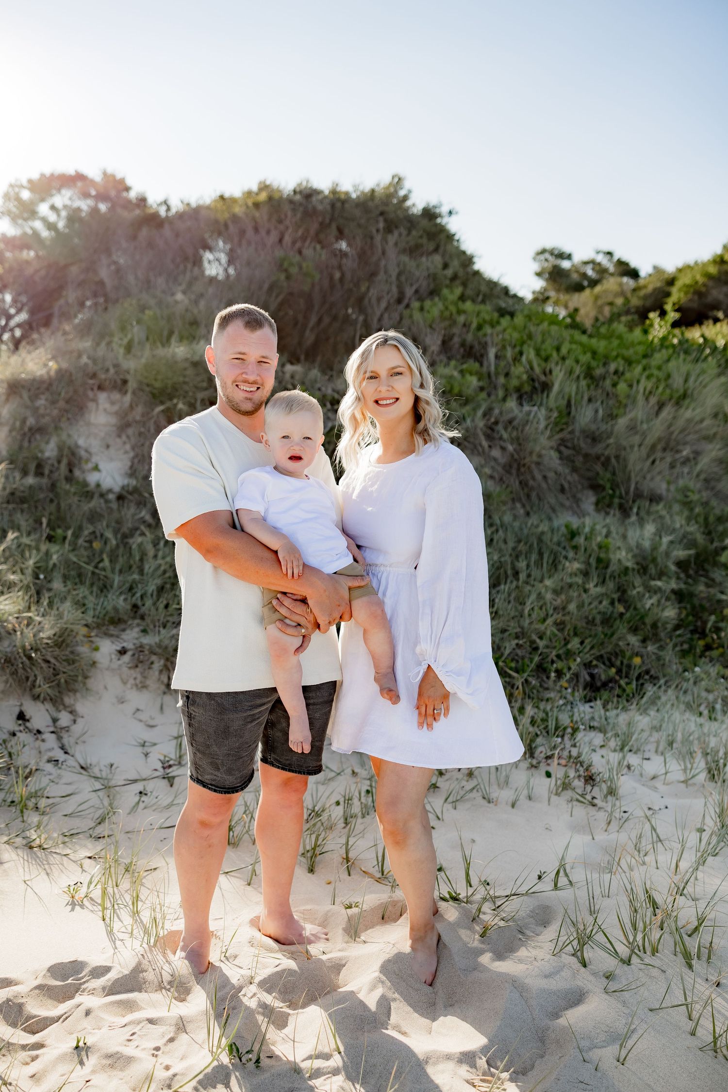 Family of three poses for portrait on sandy beach dunes at sunset.